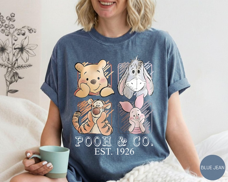 May include: Blue jean t-shirt featuring Winnie the Pooh and friends. The design includes Pooh Bear, Eeyore, Tigger, and Piglet, with the text "POOH & CO. EST. 1926". The shirt has rolled-up sleeves.