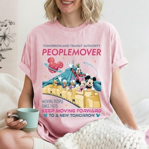 May include: Pink t-shirt featuring a Tomorrowland Transit Authority Peoplemover design. The graphic includes Mickey Mouse, Goofy, Donald Duck, and Daisy Duck. Text reads "Moving People Since 1975" and "Keep Moving Forward to a New Tomorrow."