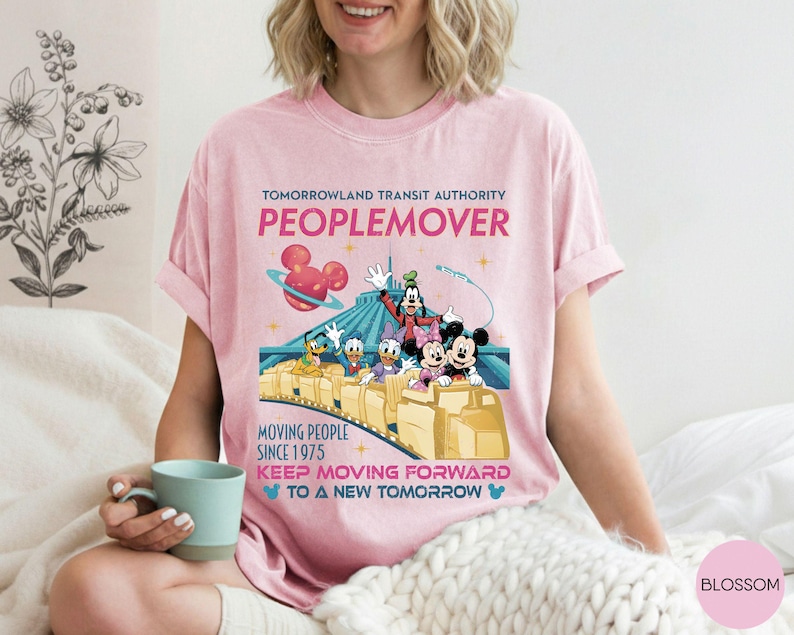 May include: Pink t-shirt featuring a Tomorrowland Transit Authority Peoplemover design. The graphic includes Mickey Mouse, Goofy, Donald Duck, and Daisy Duck. Text reads "Moving People Since 1975" and "Keep Moving Forward to a New Tomorrow."
