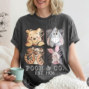 May include: Dark gray t-shirt featuring Winnie the Pooh and friends. The design includes illustrations of Pooh, Eeyore, Tigger, and Piglet, with the text "Pooh & Co. EST. 1926". The shirt is a comfortable, casual wear.