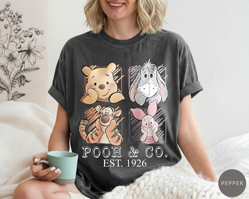 May include: Dark gray t-shirt featuring Winnie the Pooh and friends. The design includes illustrations of Pooh, Eeyore, Tigger, and Piglet, with the text "Pooh & Co. EST. 1926". The shirt is a comfortable, casual wear.