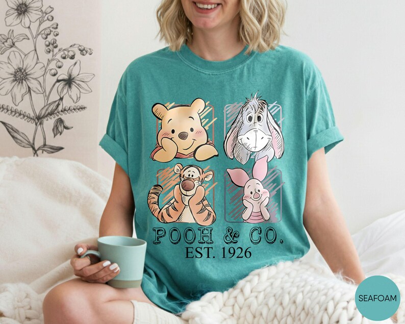 May include: Teal t-shirt featuring Winnie the Pooh, Eeyore, Tigger, and Piglet. The graphic includes the text "POOH & CO. EST. 1926". The shirt has short sleeves and a crew neck.