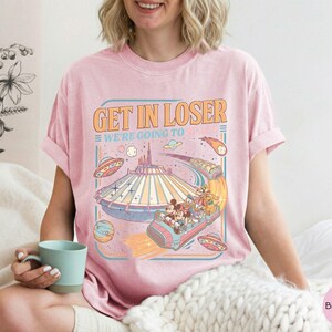 Pode incluir: T-shirt rosa claro com o texto "GET IN LOSER WE'RE GOING TO" e um gr&aacute;fico retr&ocirc; de um passeio de parque tem&aacute;tico com personagens de desenhos animados, planetas e discos voadores. A camisa tem mangas curtas e gola redonda.