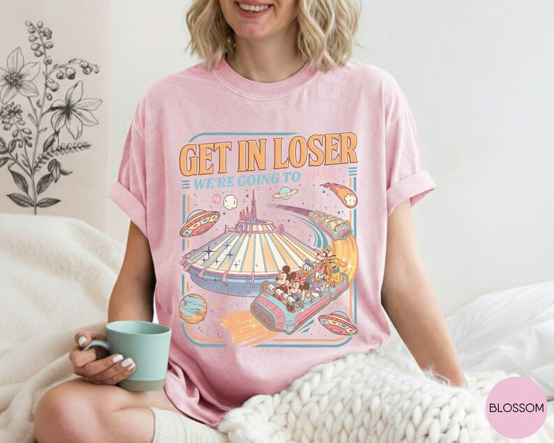 Pode incluir: T-shirt rosa claro com o texto "GET IN LOSER WE'RE GOING TO" e um gr&aacute;fico retr&ocirc; de um passeio de parque tem&aacute;tico com personagens de desenhos animados, planetas e discos voadores. A camisa tem mangas curtas e gola redonda.