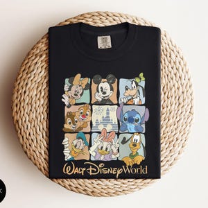 Może przedstawiać: Czarny t-shirt z kolorową siatką postaci Disneya, w tym Myszki Miki, Myszki Minnie, Goofy'ego i Stitcha. Słowa "Walt Disney World" są wydrukowane złotym pismem pod siatką. Koszulka jest złożona na tkanej powierzchni.
