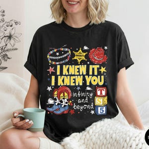 May include: A black t-shirt featuring a graphic print with the text I KNEW IT I KNEW YOU, infinity and beyond, and T S 5. The design includes a colorful beaded bracelet, a yellow sheriff star, a red cowboy hat, cow-print boots, and alphabet blocks.