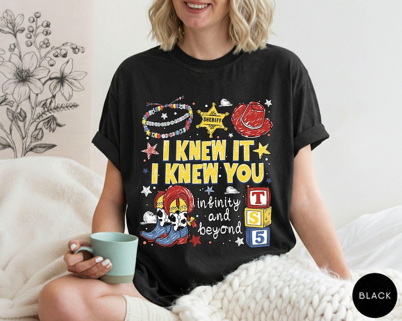 May include: A black t-shirt featuring a graphic print with the text I KNEW IT I KNEW YOU, infinity and beyond, and T S 5. The design includes a colorful beaded bracelet, a yellow sheriff star, a red cowboy hat, cow-print boots, and alphabet blocks.