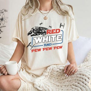 May include: An ivory t-shirt featuring a graphic print of a spaceship and small fighter crafts. The text reads RED WHITE AND PEW PEW PEW in red and white lettering with a blue outline. The design includes black line art of space vehicles.