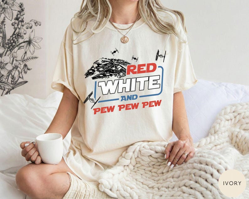 May include: An ivory t-shirt featuring a graphic print of a spaceship and small fighter crafts. The text reads RED WHITE AND PEW PEW PEW in red and white lettering with a blue outline. The design includes black line art of space vehicles.
