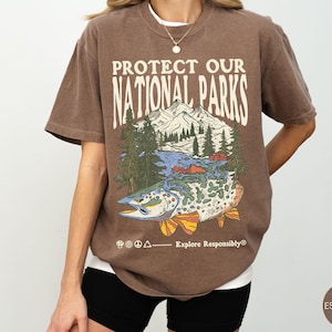 May include: Espresso-colored t-shirt with the text "PROTECT OUR NATIONAL PARKS" above a graphic of a mountain range, trees, and a fish. The phrase "Explore Responsibly" is printed below the graphic. The shirt is a relaxed fit.