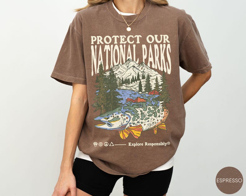 May include: Espresso-colored t-shirt with the text "PROTECT OUR NATIONAL PARKS" above a graphic of a mountain range, trees, and a fish. The phrase "Explore Responsibly" is printed below the graphic. The shirt is a relaxed fit.