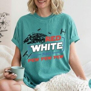 May include: A seafoam green short-sleeve t-shirt featuring a graphic print of a spaceship and TIE fighters. The text reads RED WHITE AND PEW PEW PEW in red, white, and blue lettering. The shirt has a crew neckline and a relaxed, casual fit.