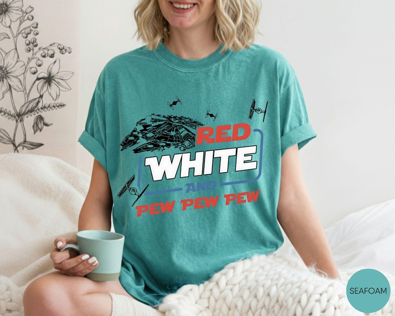 May include: A seafoam green short-sleeve t-shirt featuring a graphic print of a spaceship and TIE fighters. The text reads RED WHITE AND PEW PEW PEW in red, white, and blue lettering. The shirt has a crew neckline and a relaxed, casual fit.