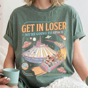 Pode incluir: T-shirt verde musgo com um gr&aacute;fico colorido que diz "GET IN LOSER WE'RE GOING TO SPACE". O gr&aacute;fico apresenta um passeio de parque tem&aacute;tico espacial com Mickey Mouse, Minnie, Pato Donald e Pateta. A camisa tem um ajuste descontra&iacute;do.