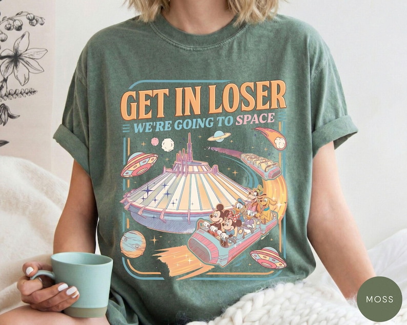 Pode incluir: T-shirt verde musgo com um gr&aacute;fico colorido que diz "GET IN LOSER WE'RE GOING TO SPACE". O gr&aacute;fico apresenta um passeio de parque tem&aacute;tico espacial com Mickey Mouse, Minnie, Pato Donald e Pateta. A camisa tem um ajuste descontra&iacute;do.