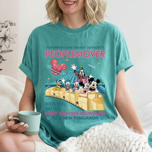 May include: Seafoam green t-shirt featuring a graphic of the Tomorrowland Transit Authority PeopleMover ride with Mickey Mouse, Minnie Mouse, Donald Duck, Daisy Duck, and Goofy. The shirt has the text "Moving People Since 1975" and "Keep Moving Forward to a New Tomorrow".