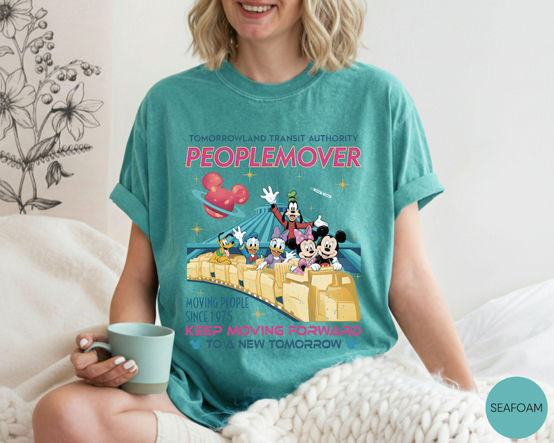May include: Seafoam green t-shirt featuring a graphic of the Tomorrowland Transit Authority PeopleMover ride with Mickey Mouse, Minnie Mouse, Donald Duck, Daisy Duck, and Goofy. The shirt has the text "Moving People Since 1975" and "Keep Moving Forward to a New Tomorrow".