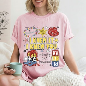 May include: A light pink t-shirt featuring a graphic print with the text I KNEW IT I KNEW YOU infinity and beyond. The design includes a colorful beaded bracelet, a yellow sheriff star, a red cowboy hat, cow-print boots, and three stacked blocks.
