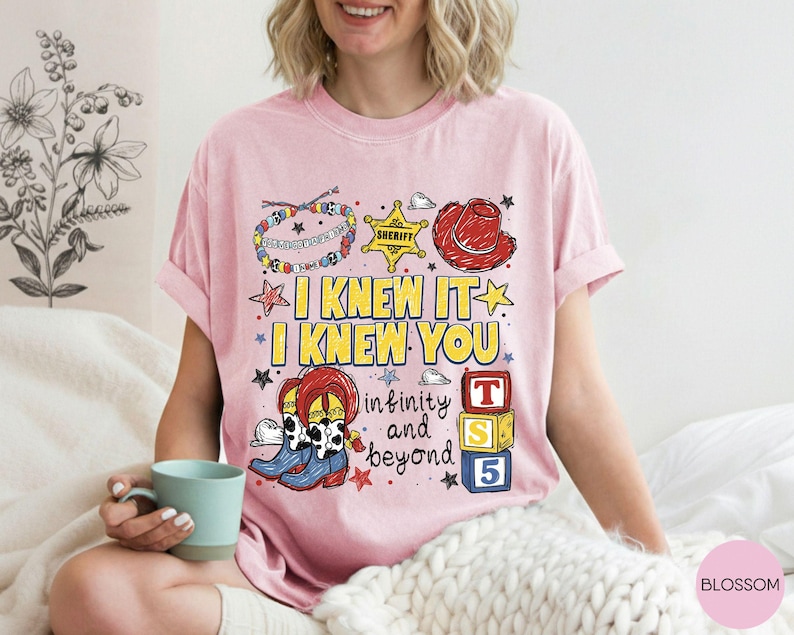 May include: A light pink t-shirt featuring a graphic print with the text I KNEW IT I KNEW YOU infinity and beyond. The design includes a colorful beaded bracelet, a yellow sheriff star, a red cowboy hat, cow-print boots, and three stacked blocks.