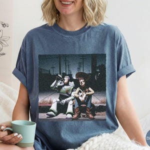 May include: A blue denim-colored t-shirt featuring a graphic print of two characters sitting on a curb at night. One character is a space ranger in a white and green suit, and the other is a cowboy in a brown hat, blue shirt, and vest.