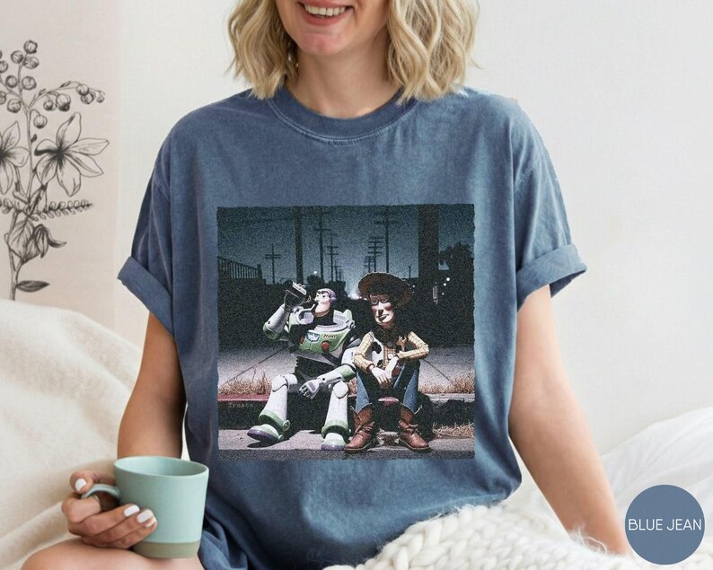 May include: A blue denim-colored t-shirt featuring a graphic print of two characters sitting on a curb at night. One character is a space ranger in a white and green suit, and the other is a cowboy in a brown hat, blue shirt, and vest.