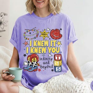 May include: A violet short-sleeve t-shirt featuring a graphic print with the text I KNEW IT I KNEW YOU infinity and beyond. The design includes a colorful beaded bracelet, a yellow sheriff star, a red cowboy hat, cowboy boots, and blocks.
