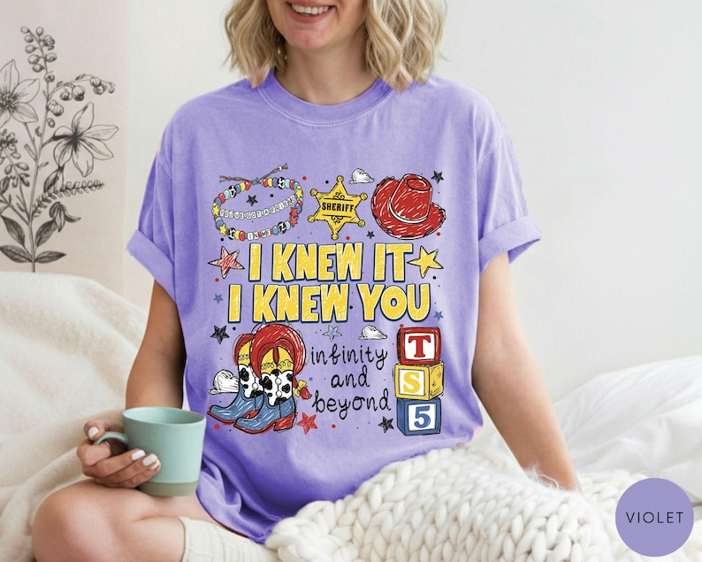 May include: A violet short-sleeve t-shirt featuring a graphic print with the text I KNEW IT I KNEW YOU infinity and beyond. The design includes a colorful beaded bracelet, a yellow sheriff star, a red cowboy hat, cowboy boots, and blocks.