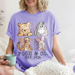 May include: A violet t-shirt featuring four cartoon characters: Winnie the Pooh, Eeyore, Tigger, and Piglet. The shirt has the text "POOH & CO. EST. 1926" printed below the characters. The shirt is a comfortable, casual style.