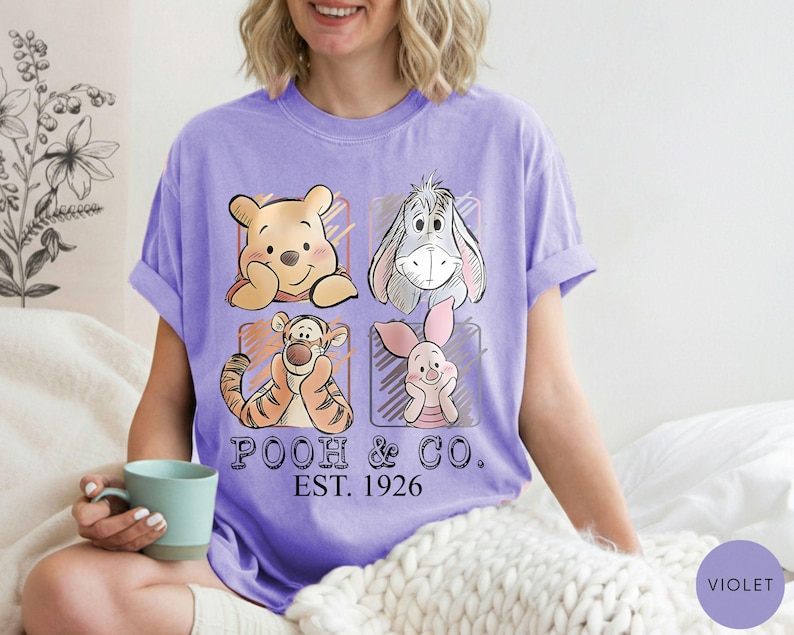May include: A violet t-shirt featuring four cartoon characters: Winnie the Pooh, Eeyore, Tigger, and Piglet. The shirt has the text "POOH & CO. EST. 1926" printed below the characters. The shirt is a comfortable, casual style.