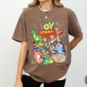 May include: A brown short-sleeve t-shirt featuring a colourful graphic print of Toy Story characters including Woody, Buzz Lightyear, Jessie, Rex, and others. The design includes the Toy Story logo and scribbled star accents in a crayon-like style.