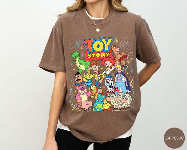 May include: A brown short-sleeve t-shirt featuring a colourful graphic print of Toy Story characters including Woody, Buzz Lightyear, Jessie, Rex, and others. The design includes the Toy Story logo and scribbled star accents in a crayon-like style.