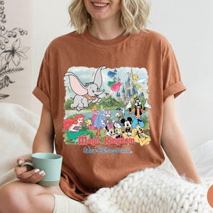 May include: A rust-colored short-sleeve t-shirt featuring a graphic print of various Disney characters including Dumbo, Ariel, Mickey Mouse, and Goofy in front of a castle, with the text Magic Kingdom Walt Disney World printed below the characters.