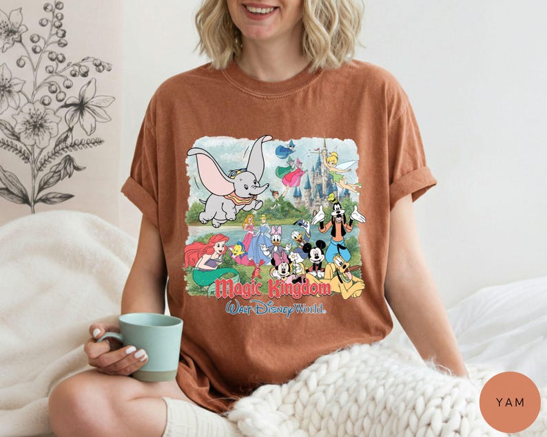 May include: A rust-colored short-sleeve t-shirt featuring a graphic print of various Disney characters including Dumbo, Ariel, Mickey Mouse, and Goofy in front of a castle, with the text Magic Kingdom Walt Disney World printed below the characters.