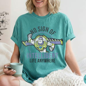 Puede incluir: Camiseta verde mar con un dibujo animado de Buzz Lightyear y el texto "NO SIGN OF INTELLIGENT LIFE ANYWHERE". El dise&ntilde;o presenta colores verdes, morados y blancos. Es una prenda informal.