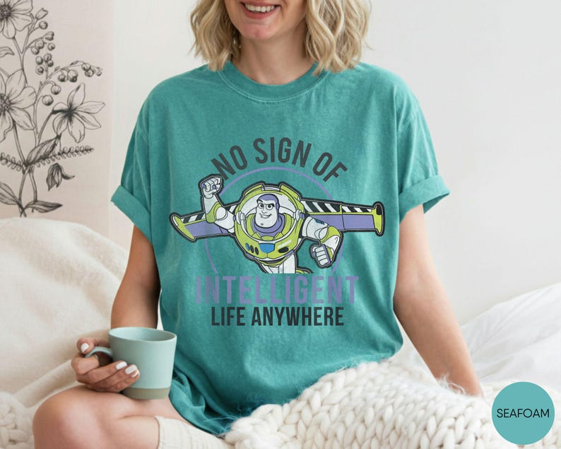 Puede incluir: Camiseta verde mar con un dibujo animado de Buzz Lightyear y el texto "NO SIGN OF INTELLIGENT LIFE ANYWHERE". El dise&ntilde;o presenta colores verdes, morados y blancos. Es una prenda informal.