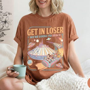 Pode incluir: Camiseta marrom com o texto "GET IN LOSER WE'RE GOING TO SPACE" e um gr&aacute;fico colorido de um passeio tem&aacute;tico espacial. O gr&aacute;fico inclui personagens de desenhos animados e discos voadores.