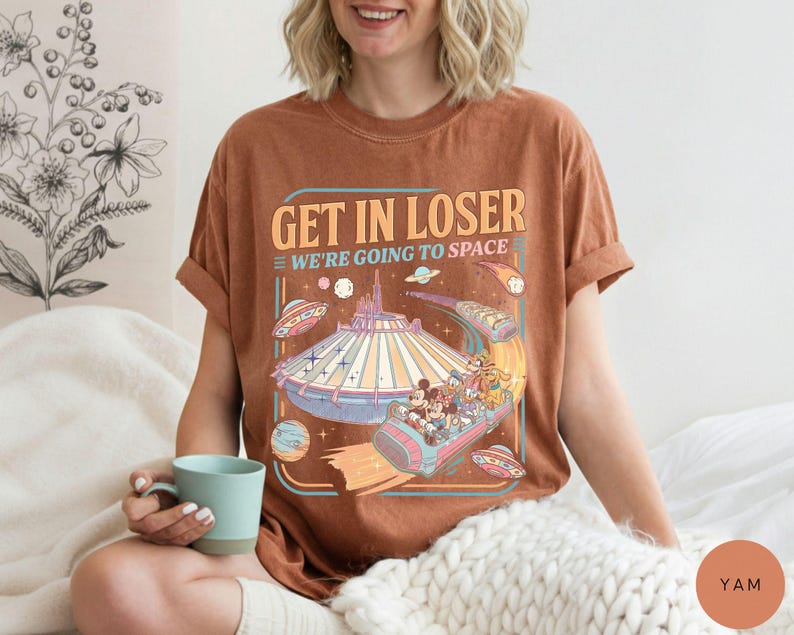 Pode incluir: Camiseta marrom com o texto "GET IN LOSER WE'RE GOING TO SPACE" e um gr&aacute;fico colorido de um passeio tem&aacute;tico espacial. O gr&aacute;fico inclui personagens de desenhos animados e discos voadores.