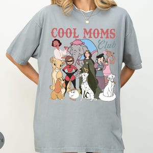 May include: A light gray t-shirt with the text "COOL MOMS Club" in red and blue. The shirt features a graphic of cartoon characters, including a lion cub, a dog, and a cat.