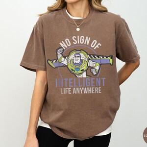 Puede incluir: Camiseta marr&oacute;n con un gr&aacute;fico de Buzz Lightyear y el texto "NO SIGN OF INTELLIGENT LIFE ANYWHERE". El dise&ntilde;o incluye un marco circular alrededor del personaje. La camiseta tiene un corte holgado y mangas cortas.