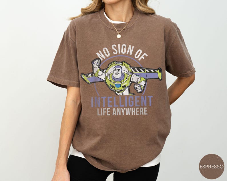 Puede incluir: Camiseta marr&oacute;n con un gr&aacute;fico de Buzz Lightyear y el texto "NO SIGN OF INTELLIGENT LIFE ANYWHERE". El dise&ntilde;o incluye un marco circular alrededor del personaje. La camiseta tiene un corte holgado y mangas cortas.