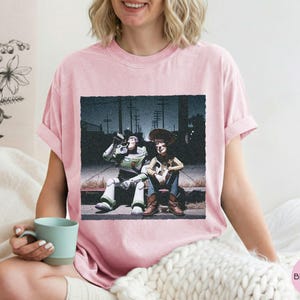 May include: A light pink short-sleeve t-shirt featuring a graphic print of two characters sitting on a curb at night. One character is a white, green, and purple space ranger, and the other is a cowboy wearing a brown hat, vest, and blue jeans.