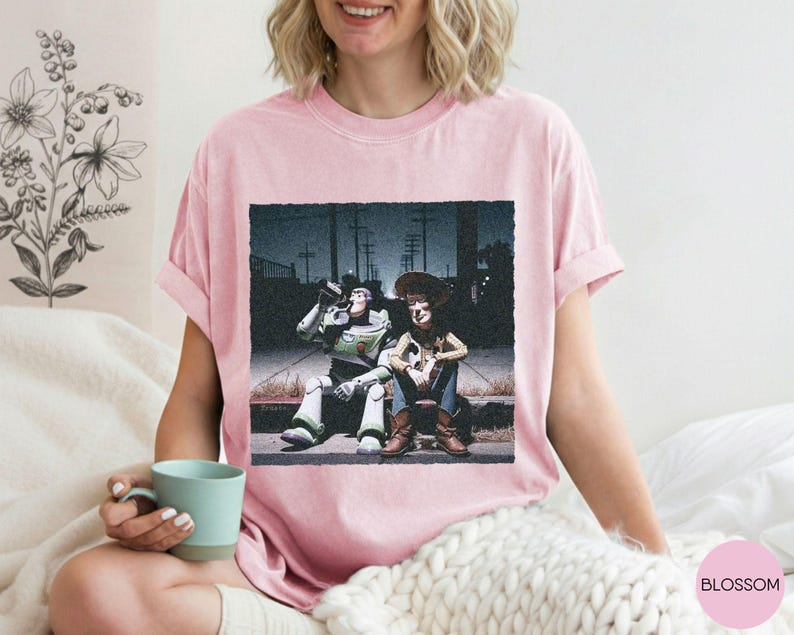 May include: A light pink short-sleeve t-shirt featuring a graphic print of two characters sitting on a curb at night. One character is a white, green, and purple space ranger, and the other is a cowboy wearing a brown hat, vest, and blue jeans.
