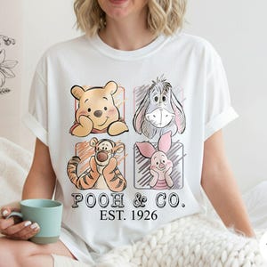 May include: White t-shirt featuring four cartoon character portraits: Winnie the Pooh, Eeyore, Tigger, and Piglet. The shirt has the text "POOH & CO. EST. 1926" printed below the characters. The shirt is being worn.
