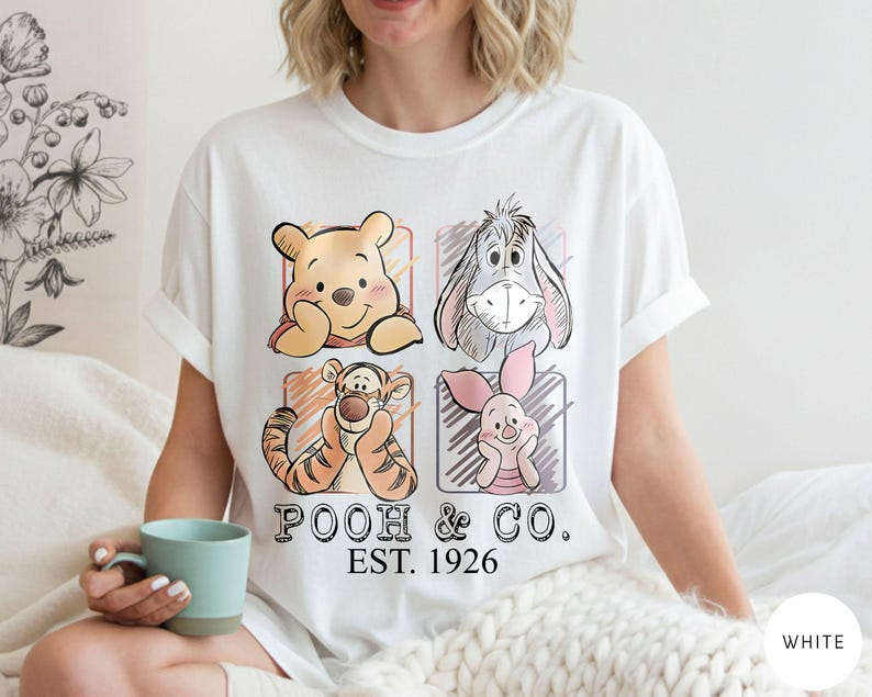 May include: White t-shirt featuring four cartoon character portraits: Winnie the Pooh, Eeyore, Tigger, and Piglet. The shirt has the text "POOH & CO. EST. 1926" printed below the characters. The shirt is being worn.