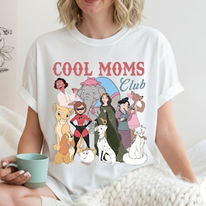 May include: White t-shirt with the text "COOL MOMS Club" in pink and red. The shirt features a cartoon graphic of various characters. The shirt is made of a soft material.