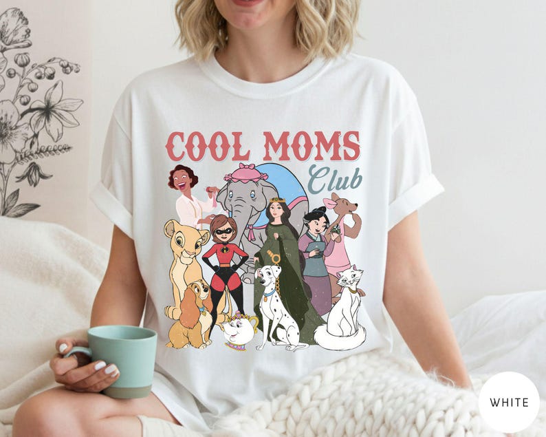 May include: White t-shirt with the text "COOL MOMS Club" in pink and red. The shirt features a cartoon graphic of various characters. The shirt is made of a soft material.