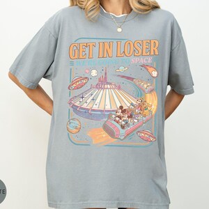 Pode incluir: Camiseta cinza claro com um gr&aacute;fico de um passeio tem&aacute;tico espacial. O design inclui o texto "GET IN LOSER WE'RE GOING TO SPACE" em laranja e azul, juntamente com personagens de desenhos animados e planetas.