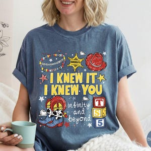 May include: A blue denim-colored t-shirt featuring a graphic print with the text I KNEW IT I KNEW YOU and infinity and beyond. The design includes a sheriff badge, a red cowboy hat, cowboy boots, a beaded bracelet, and three colorful toy blocks.