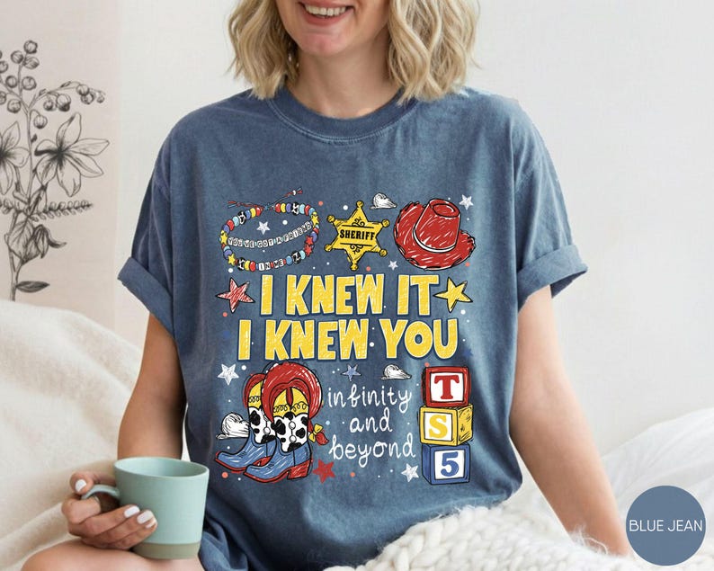 May include: A blue denim-colored t-shirt featuring a graphic print with the text I KNEW IT I KNEW YOU and infinity and beyond. The design includes a sheriff badge, a red cowboy hat, cowboy boots, a beaded bracelet, and three colorful toy blocks.