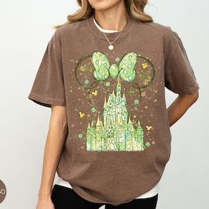 K&ouml;nnte beinhalten: Espresso-farbenes T-Shirt mit einem gr&uuml;nen Schloss-Design, Schleife und Mickey Mouse-Symbolen. Die Grafik verwendet Gr&uuml;n- und Gelbt&ouml;ne mit einem Vintage-Look. Das Shirt hat kurze &Auml;rmel und eine lockere Passform.